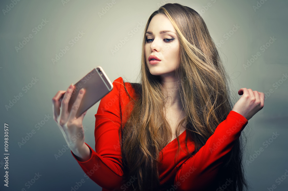 Naklejka premium Beautiful woman in red dress with perfect cute face and blue eyes taking selfie with smartphone in studio.