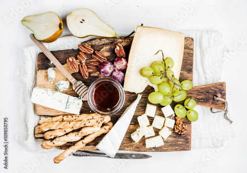 Cheese appetizer set. Various types of cheese, honey, grapes, pear, nuts and bread grissini sticks on rustic wooden board over white background photo