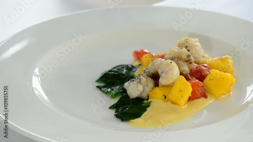 Chef adding pieces of fish and asparagus on a plate with sauce, spinachs, tomatoes and mango photo