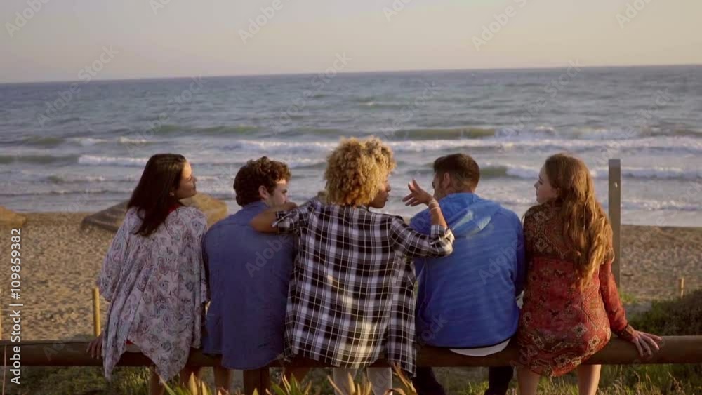 Back view of young people while sitting on wooden fence and enjoy leisure time on the beach at the evening.