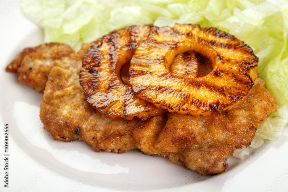 Fried schnitzel ( tenderized and breaded pork fillet cutlet ) with grilled pineapple slices and lettuce in background