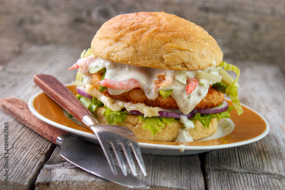 Salmon and shrimp burger , mayonnaise on bottom bun , breaded salmon shrimp patty and tartar sauce with fresh shrimps on gray wooden boards background