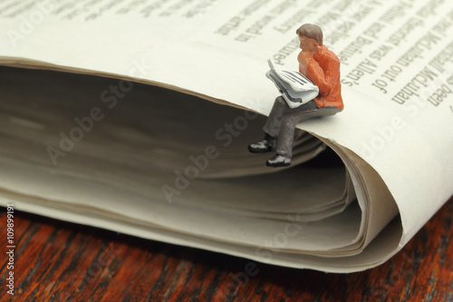 Miniature figure of a man reading a newspaper while sitting on a full size newspaper