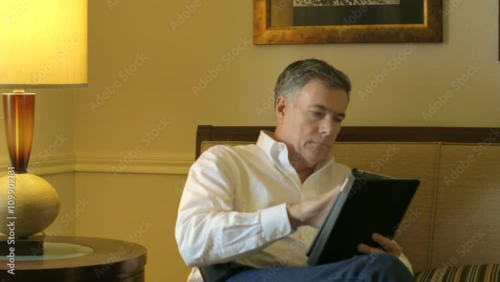 A handsome mature man sitting alone in his living room using an electronic tablet. A handsome mature man sitting alone in his living room using an electronic tablet.