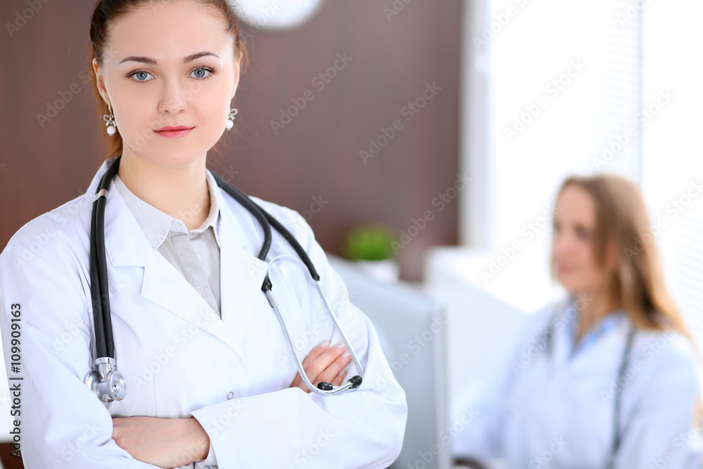 Beautiful young smiling female doctor standing in a hospital with her colleague in the background Beautiful young smiling female doctor standing in a hospital with her colleague in the background