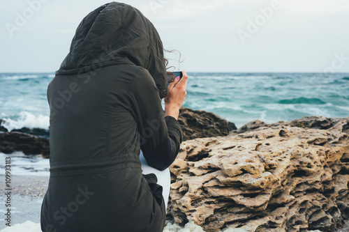 Girl in the green hoodie sitting on the beach photographing the