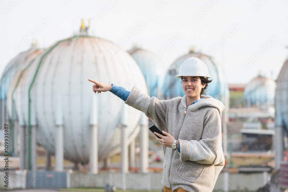 successful independent engineer woman pointing and smiling on industrial area with safety helmet and phone. Pioneer woman at work with spherical tanks. successful independent engineer woman pointing and smiling on industrial area with safety helmet and phone. Pioneer woman at work with spherical tanks.