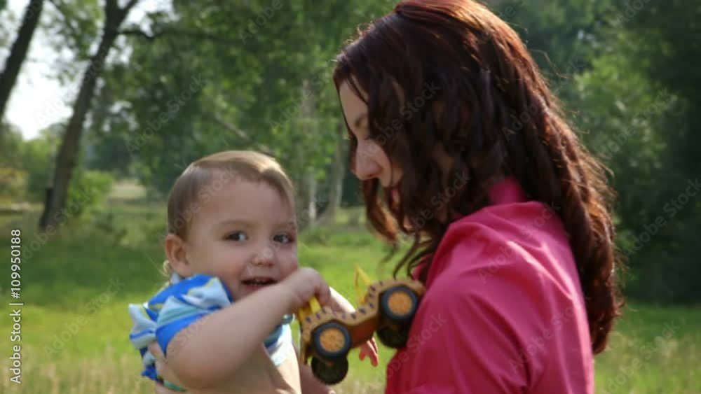 A young mother outdoors in a park area on a sunny morning encourages her little son to play with a ball. Scene moves up past wildflowers to action with mother and son. A young mother outdoors in a park area on a sunny morning encourages her little son to play with a ball. Scene moves up past wildflowers to action with mother and son.