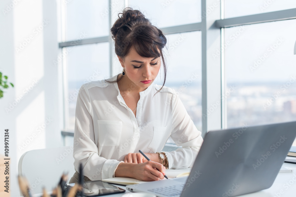Female writer working in light modern office, writing down new ideas her notebook, searching information using portable computer at workplace