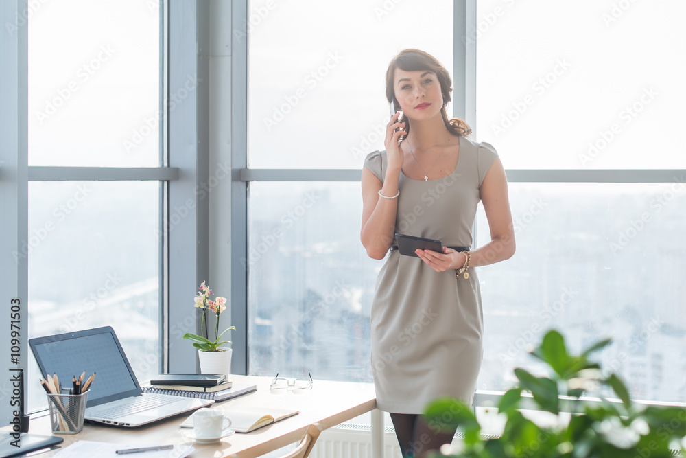 Young female consultant having business call in office at her workplace. Secretary speaking on the smart phone, holding pda. Young female consultant having business call in office at her workplace. Secretary speaking on the smart phone, holding pda.