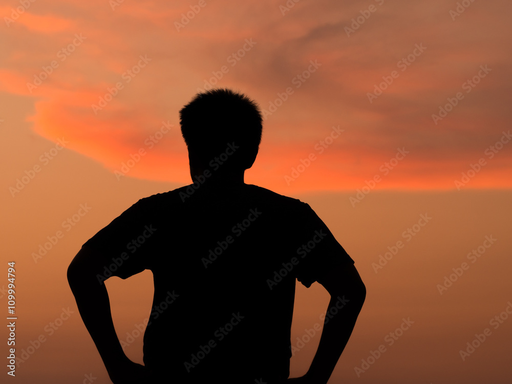 Silhouette man standing with arms akimbo at sunset orange sky in background.