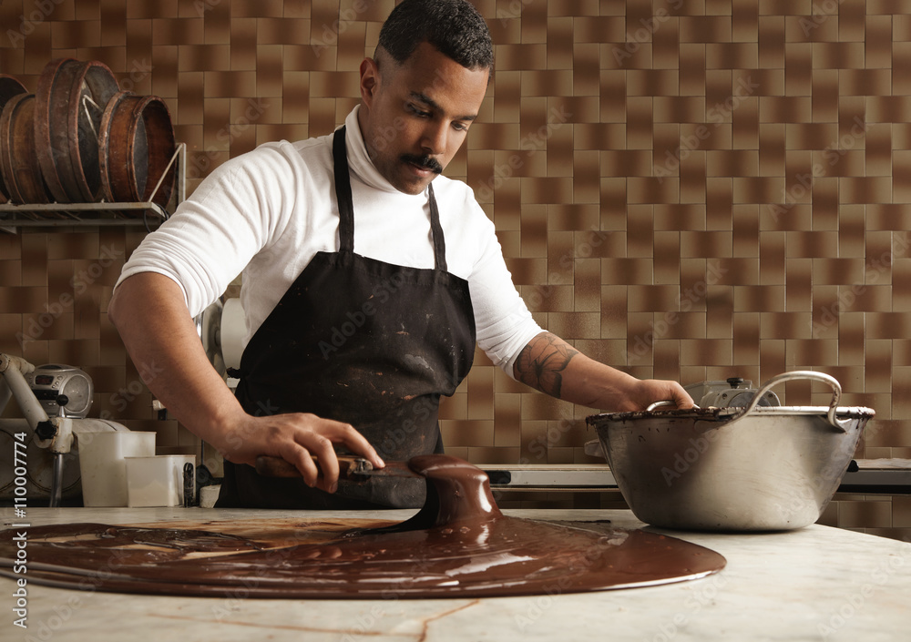 Black man professional chief collects tasty melted chocolate from vintage  marble table to big steel pot in his artisan rustic kitchen with industrial  retro machines. Mirrored in chocolate on table. Photos