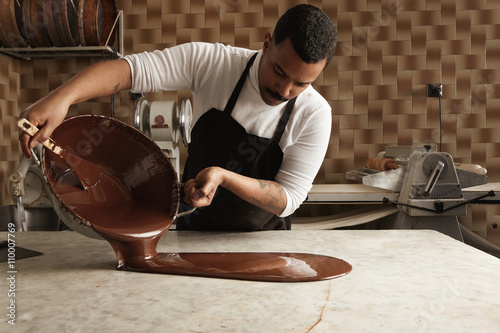 Картината върху платно Black man professional chief pours tasty melted chocolate from one big steel pot to vintage marble table in his artisan rustic kitchen with industrial retro machines