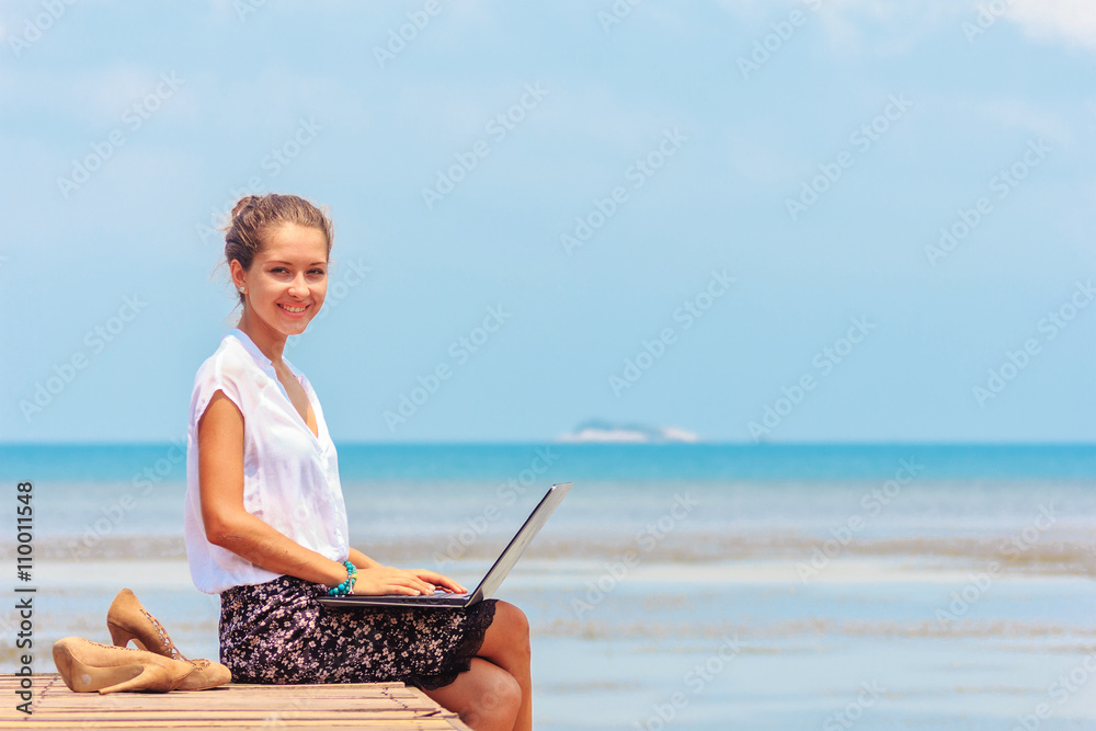 Young woman sitting with a laptop on the wooden platform edge at the sea. Lady freelancer in office suit working outdoor. Looking at camera and smiling.