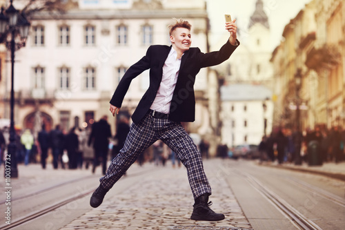 Full body portrait of young happy smiling handsome man running, jumping & taking selfie photo on old street. City lifestyle. Old european architecture on background. Toned style instagram filters
 photo