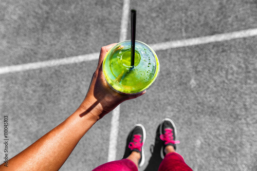 Green smoothie woman drinking plastic cup breakfast meal takeaway to go after morning run on city streets. Healthy lifestyle sporty person pov of hand holding glass with running shoes feet selfie.