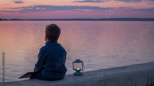 child reading a book with lantern at sunset near water