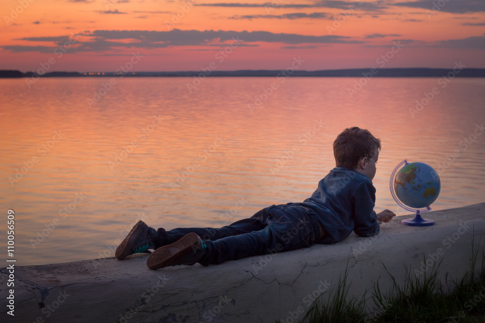 Child lying and looking at the globe during sunset near water