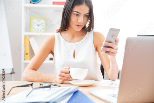 Businesswoman holding cup with coffee and using smartphone