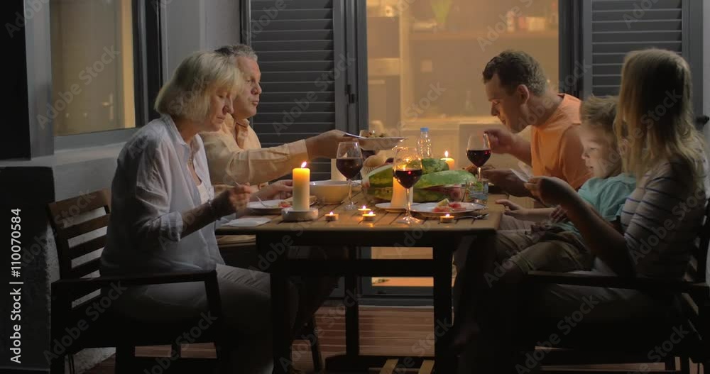 Cheerful family enjoying meal and wine during evening dinner outdoor. Little boy playing tablet computer on mothers knees Cheerful family enjoying meal and wine during evening dinner outdoor. Little boy playing tablet computer on mothers knees