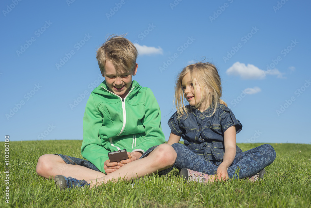 Happy blond children using smartphone (watching movie or playing game) sitting on the grass. Brother and sister. Summertime Happy blond children using smartphone (watching movie or playing game) sitting on the grass. Brother and sister. Summertime