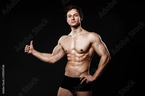 Muscular and fit young bodybuilder fitness male model showing thumb-up and posing over black background. Perfect fitness body. Ideal for commercial.