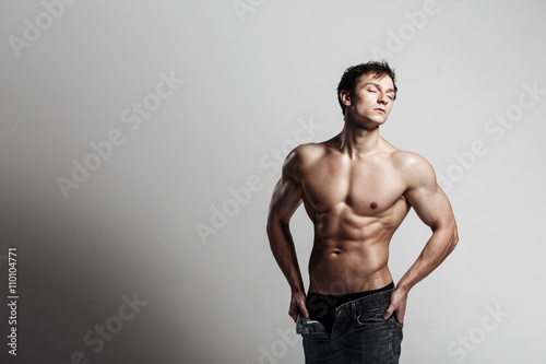 Handsome athletic man looking side in unbuttoned jeans. Strong bodybuilder with six pack  perfect abs  shoulders  biceps  triceps and chest. Great fitness body. Ideal for commercial. Gray background