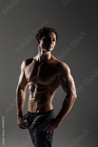 Handsome athletic man looking side in unbuttoned jeans. Strong bodybuilder with six pack, perfect abs, shoulders, biceps, triceps and chest. Great fitness body. Ideal for commercial. Gray background photo