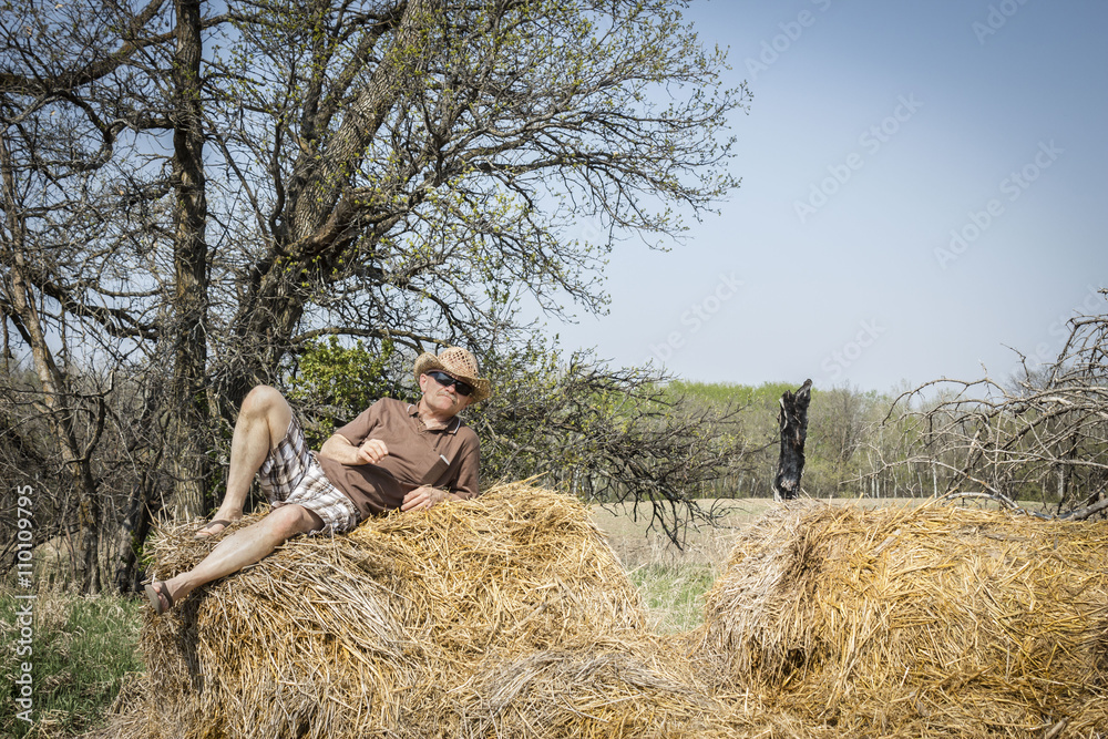 horizontal image of a man wearing cowboy hat leaning back on an old hay bale with trees in background in the daytime in the summer. horizontal image of a man wearing cowboy hat leaning back on an old hay bale with trees in background in the daytime in the summer.