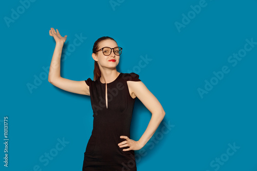 Colour studio portrait of pretty smiling happy young woman in glasses pointing upward. Blue background isolated