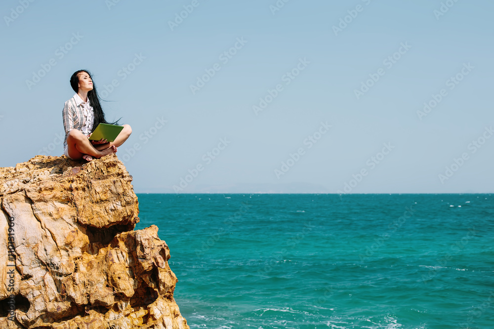 Beautiful business woman sitting on stone cliff on blue sea beach, enjoy freedom, using laptop computer