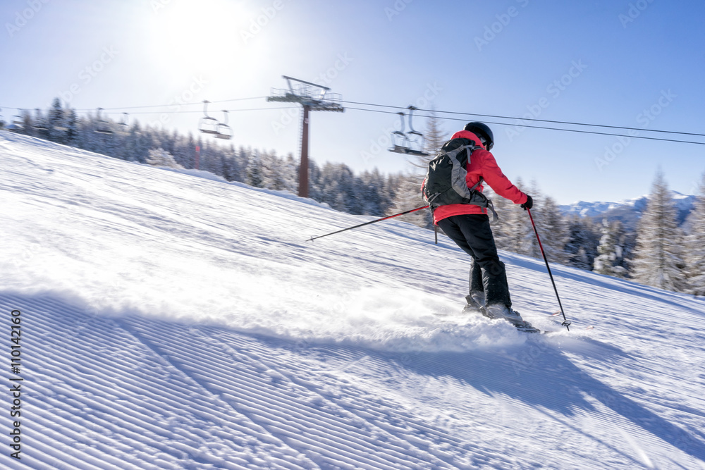 Female skiier dressed in red jacket enjoys slopes in Italy with sun behind her.