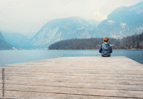 Little boy sits on the wooden pier near the blue mountain lake
