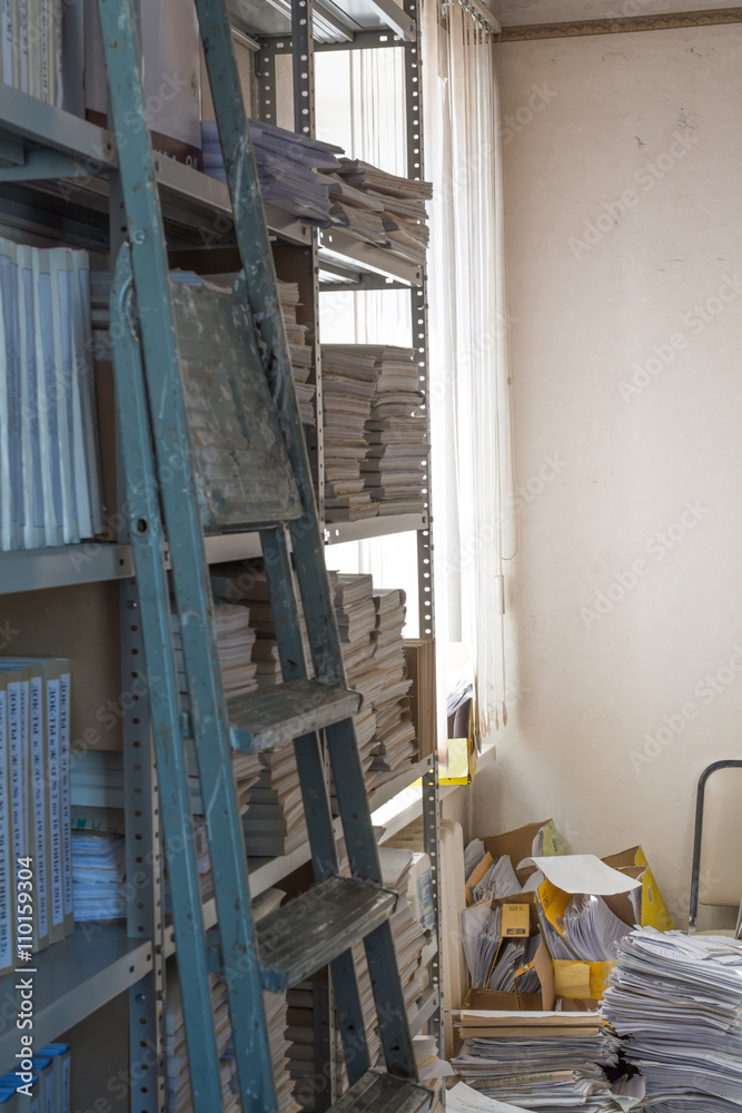 A vertical image of archive in which mess. A folding ladder on foreground. Main focus on mess.