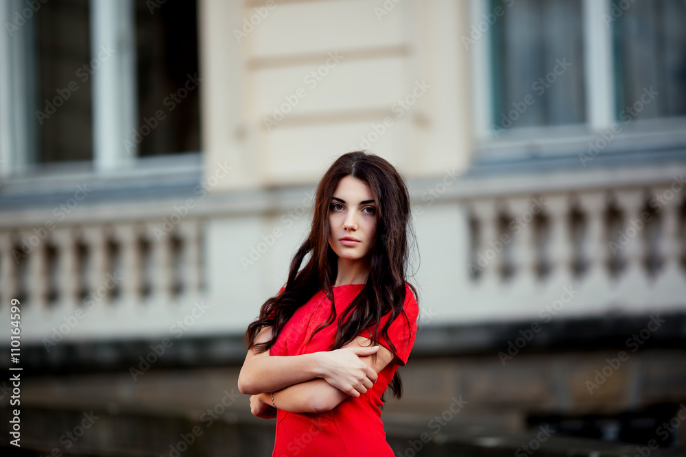 Fototapeta premium Street fashion concept: Portrait of a beautiful young woman wearing a red dress walking in the city. Old architecture background