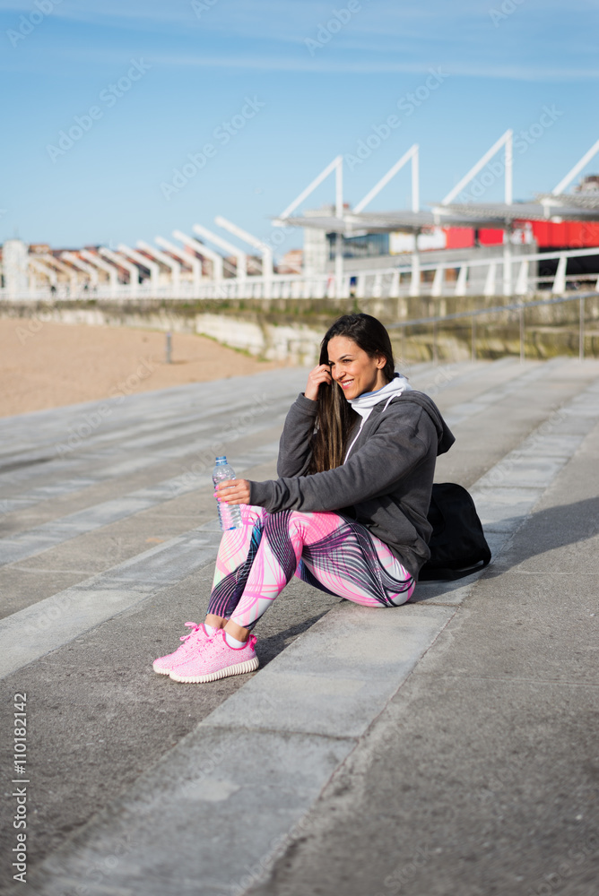 Female urban athlete sitting near the beach for resting and drinking water after outdoor workout. Sporty woman wearing fashion sport pink pattern leggings.