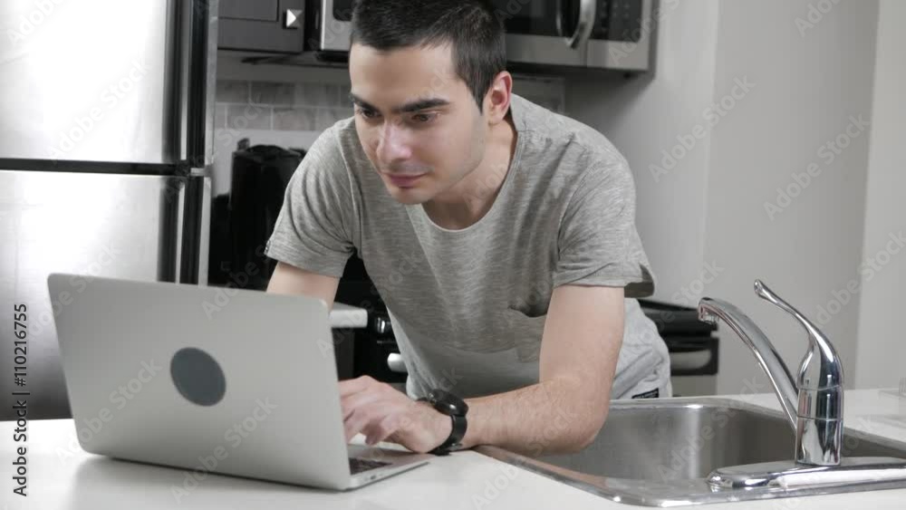 Young man in a home kitchen using a laptop computer on the counter.
