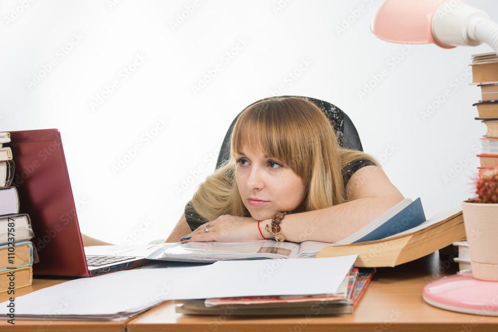 The girl at the desk crammed with books and papers sadly looks at the laptop with her head resting on his hand on the table The girl at the desk crammed with books and papers sadly looks at the laptop with her head resting on his hand on the table