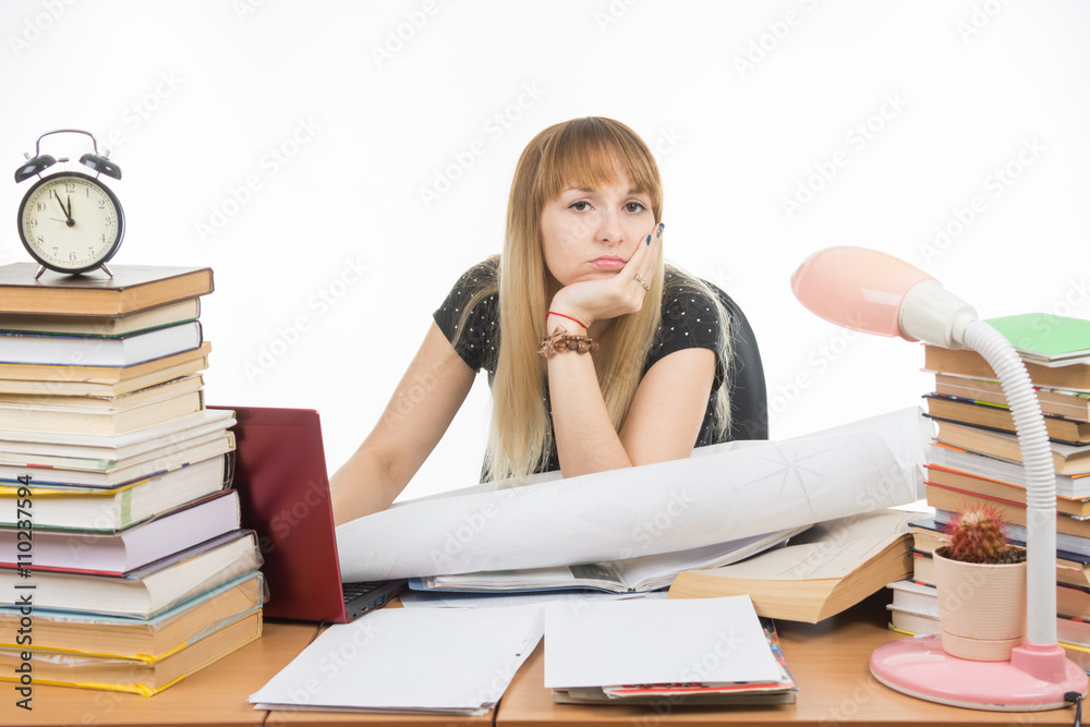 Girl student sitting sadly at the table crammed with books and papers Girl student sitting sadly at the table crammed with books and papers