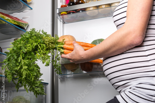 Pregnant woman opening fridge with vegetables and picking carrots