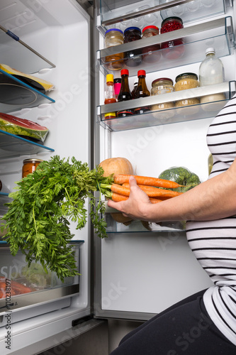 Pregnant woman opening fridge with vegetables and picking carrots