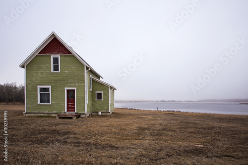 Old two storey green and red abandoned house in front of a fog shrouded lake in countryside landscape