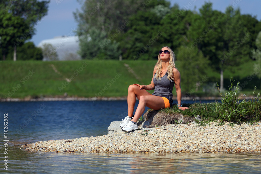 Blonde fitness girl sitting and relaxing at a river bank Blonde fitness girl sitting and relaxing at a river bank