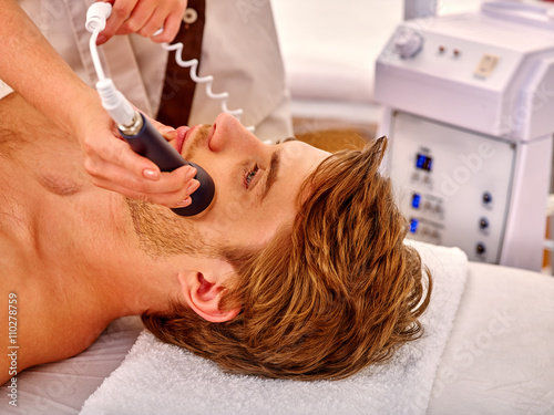 Young man luxuriating on electroporation  facial massage therapy at beauty salon. Man takes care of his face through facing massage. photo