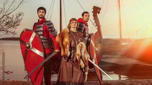 Slavic princess and two warriors with swords and shields on the warships background.