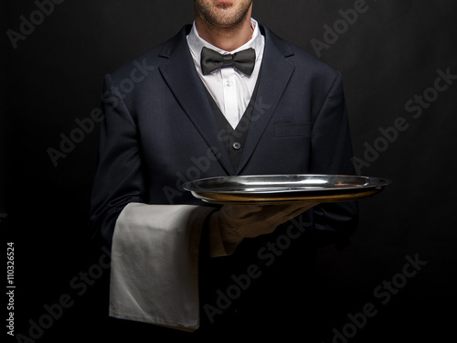 Waiter in black suit holding tray over black background.