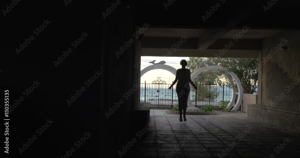 Woman skipping rope in dark yard of hotel or house. Black female silhouette in headphones against waterfront background Woman skipping rope in dark yard of hotel or house. Black female silhouette in headphones against waterfront background