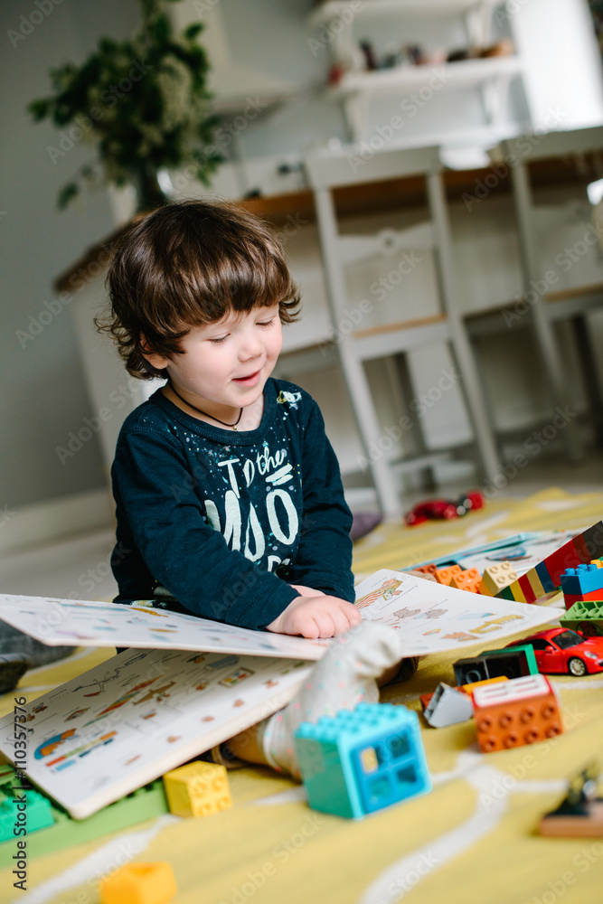 Little boy holds a book and sees the drawings. The child sits on the floor. Near scattered toys. The concept of family Little boy holds a book and sees the drawings. The child sits on the floor. Near scattered toys. The concept of family