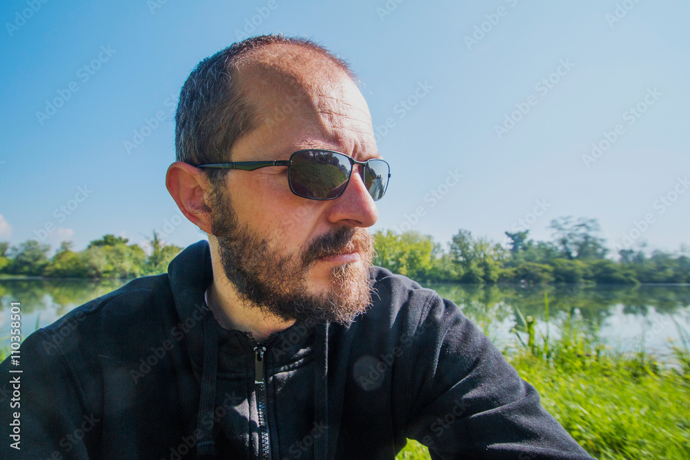Bearded and mustache man with sun glasses in black hoodie portrait in nature, serious expression, lake in the background, looking in distance, adventure concept Bearded and mustache man with sun glasses in black hoodie portrait in nature, serious expression, lake in the background, looking in distance, adventure concept