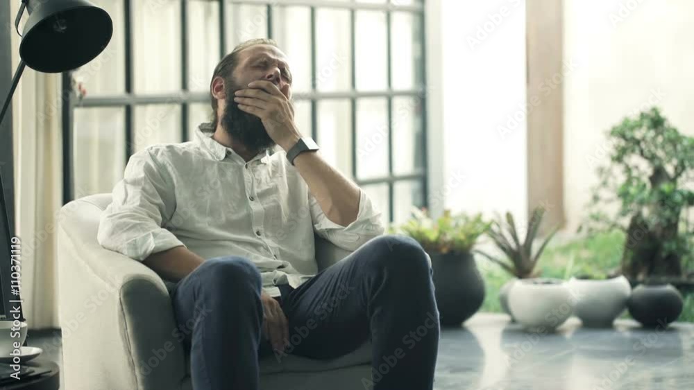 Young man yawning and falling asleep on armchair at home
